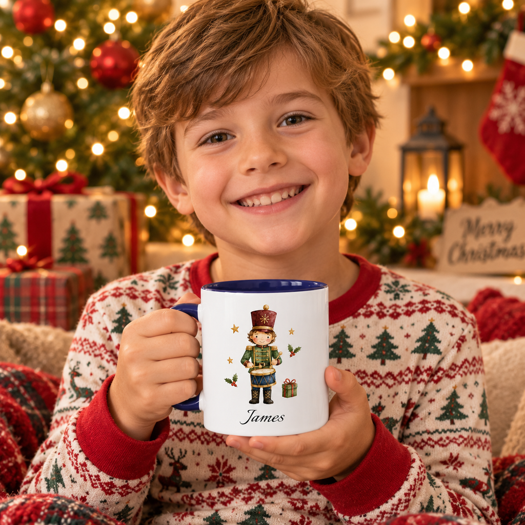 A smiling young boy in a festive Christmas sweater holds a white ceramic mug with a dark blue handle and interior. The mug is printed with a nutcracker illustration and the name 'James' at the bottom.