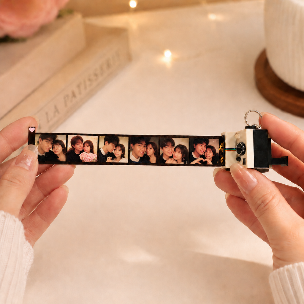 A person holds a long photo strip camera across their hands, showing a row of printed couple photos. The photo strip camera has a black and white body with a small metal keyring on the end.