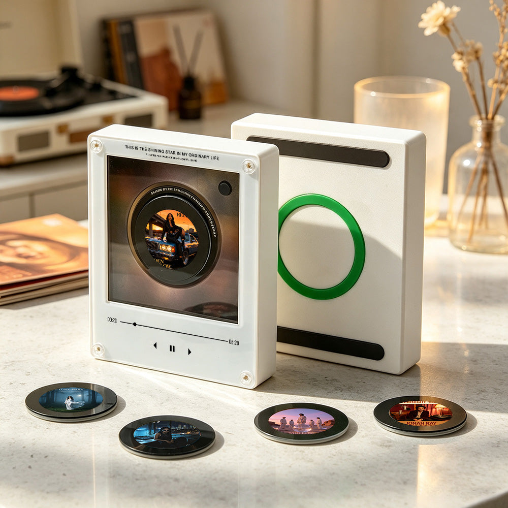 A white rectangular music player device with a circular screen showing album art, standing beside a matching white case with a green ring detail. Four small round printed discs are placed in front on a light marble surface in a styled indoor setting with books, a record player, and a vase in the background.