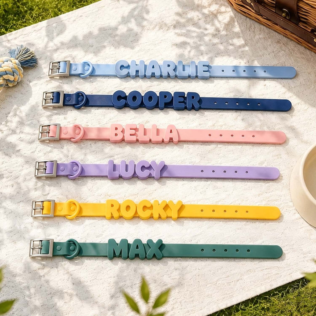 Seven personalized pet collars in different colors are laid out on a white blanket outdoors. The collars are light blue, navy blue, pink, lavender purple, yellow, and dark green, each featuring a raised name plate (CHARLIE, COOPER, BELLA, LUCY, ROCKY, MAX) and a silver metal buckle.